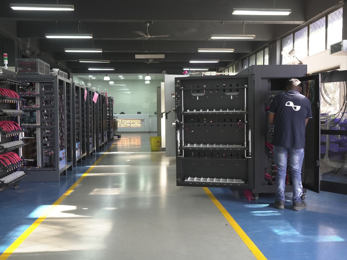 An Ador Digatron engineer connecting cables inside a battery test cabinet, with rows of test racks behind