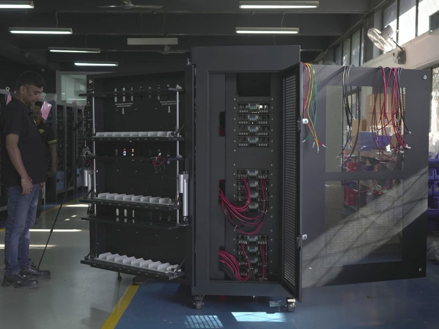 An Ador Digatron technician inspecting wiring and component trays inside an open battery test cabinet
